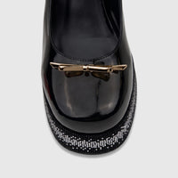 Close-up of a black patent leather Bulbul Ladies Block Heel Mary Jane shoe with a gold bow and bling platform.
