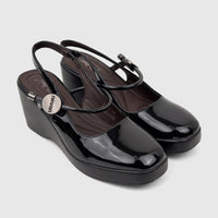 A pair of shiny black patent leather Bulbul Ladies Dress Block Heel Sandals with wedge heels and slingback straps on a white