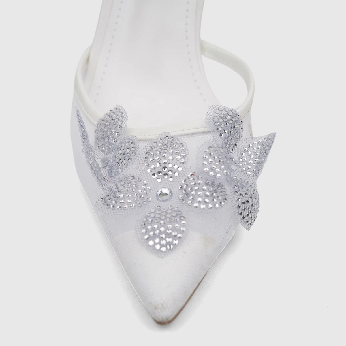 Close-up of a white pointed-toe evening heel sandal featuring a sparkling silver rhinestone flower on the front.