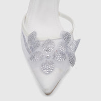 Close-up of a white pointed-toe evening heel sandal featuring a sparkling silver rhinestone flower on the front.