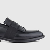 Black leather men's dress slip-on shoe with fringe detail on the vamp, viewed from a close-up angle.