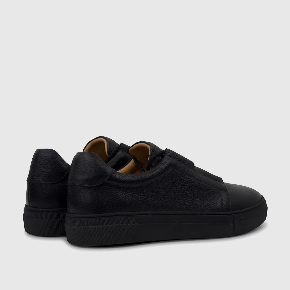 A pair of black men's leather slip-on sneakers with textured uppers and thick black soles, viewed from a rear-side angle on