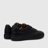 A pair of black men's leather slip-on sneakers with textured uppers and thick black soles, viewed from a rear-side angle on