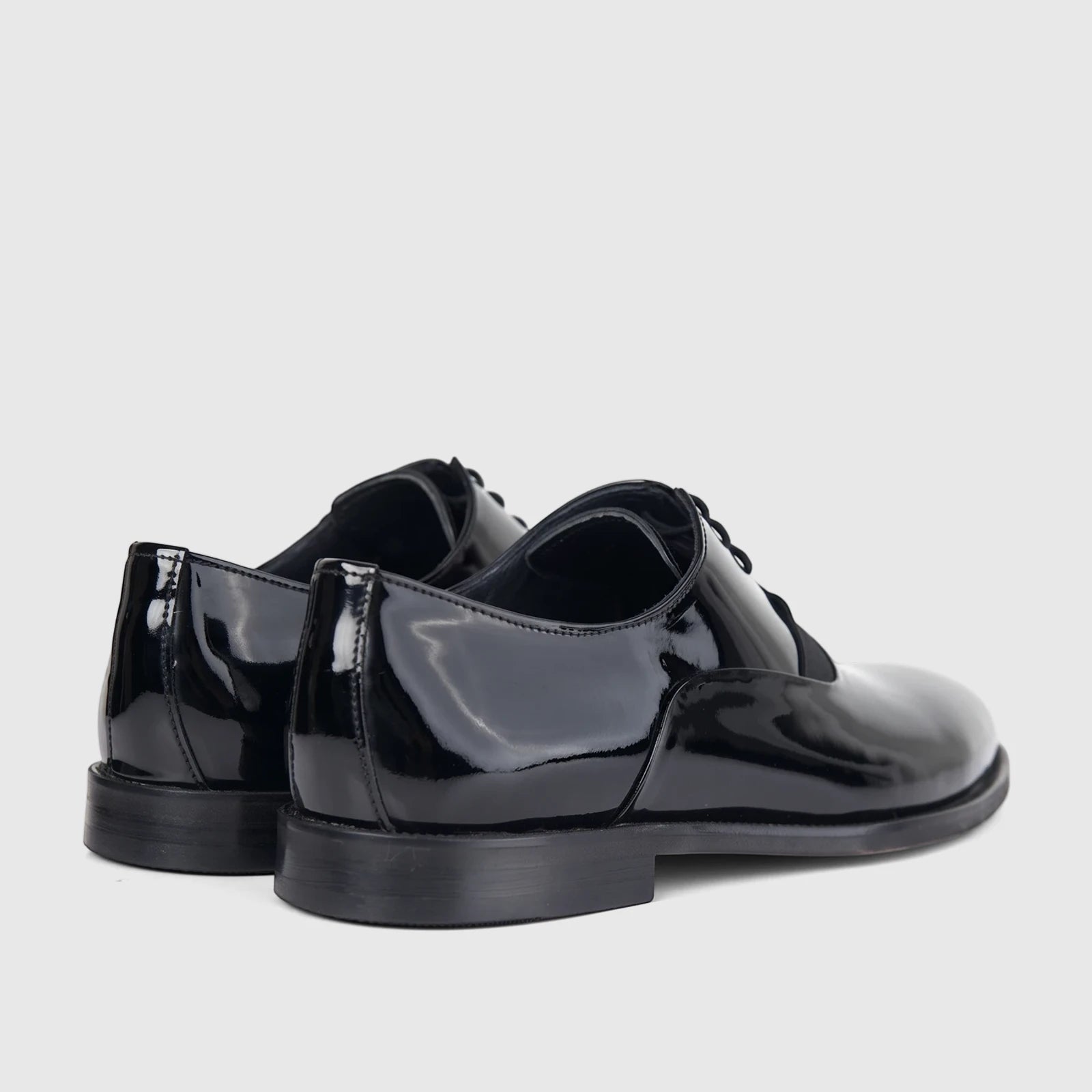 Two shiny black patent leather men's dress shoes with laces, viewed from the back-right on a a white background.