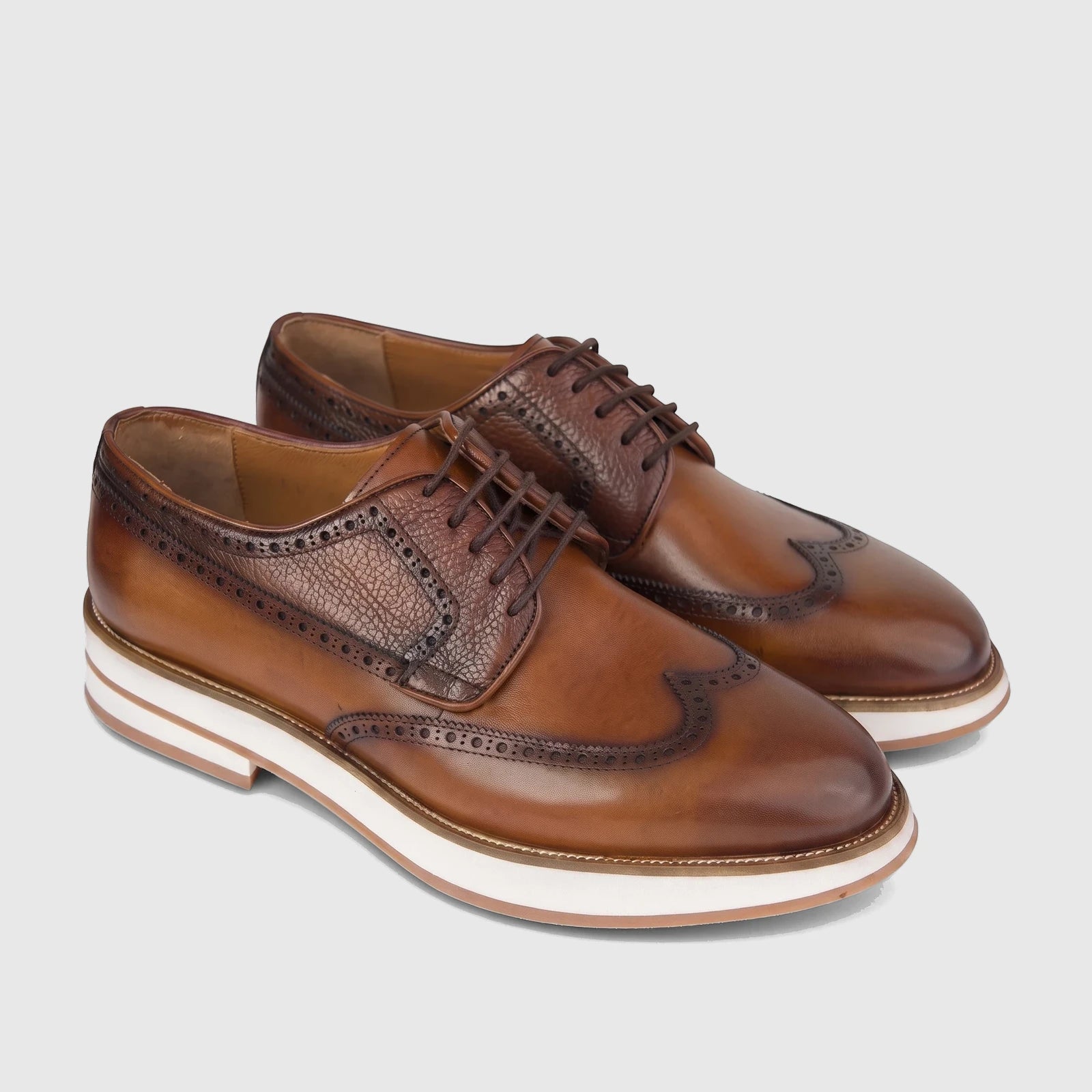 A pair of men's tan leather chunky sole lace-up brogues with wingtip details and white soles, presented at an angle.