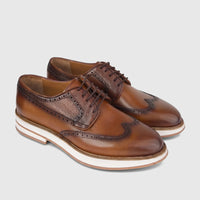 A pair of men's tan leather chunky sole lace-up brogues with wingtip details and white soles, presented at an angle.