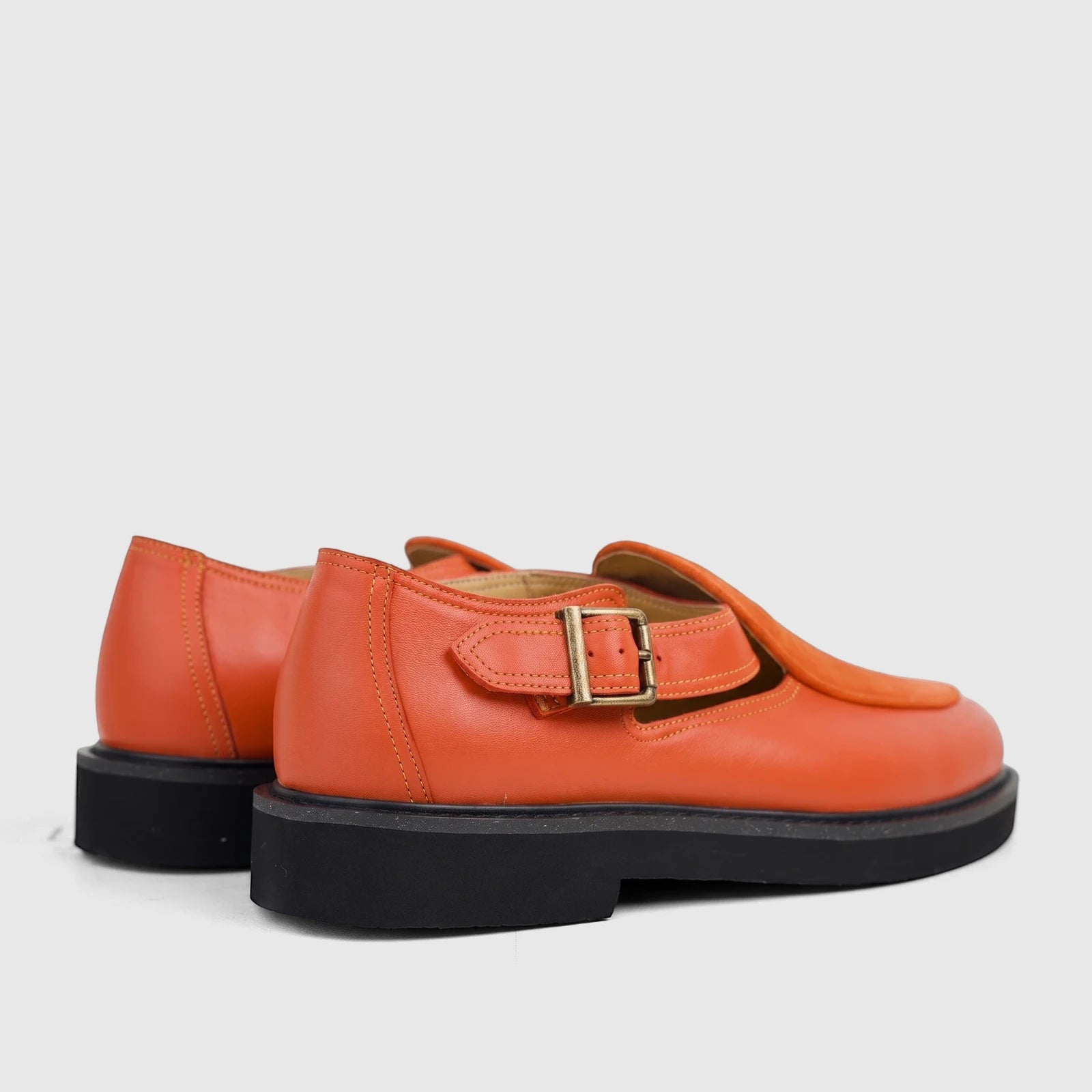 Pair of orange leather chunky sole sandals with a bronze buckle, seen from a rear-side angle on a white background.