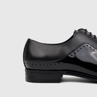 Close-up side view of a black patent leather men's lace-up dress shoe, highlighting the textured and glossy panels.