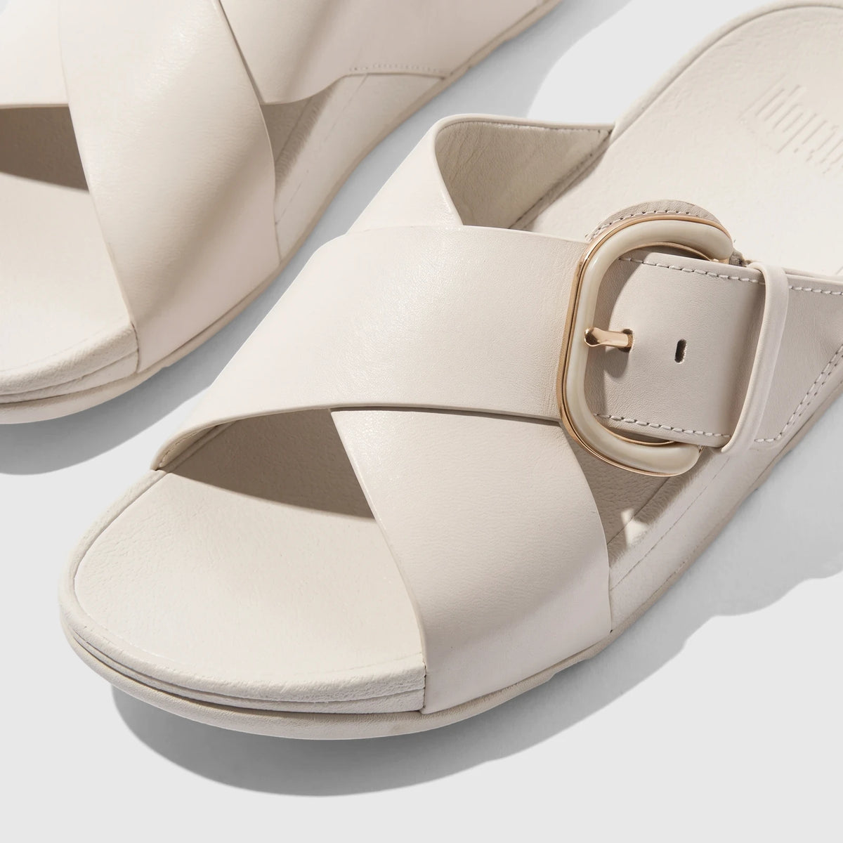 Close-up of a pair of Fit Flop Lulu Resin Buckle leather cross slide sandals in a cream color, angled on a flat surface.