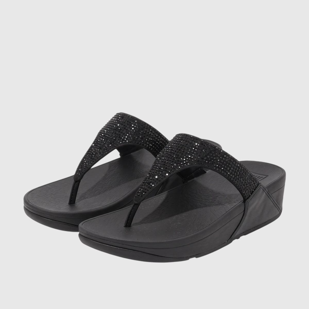 A pair of black FitFlop Lulu Crystal Embellished comfort thong sandals, featuring sparkling straps and a platform sole, on a