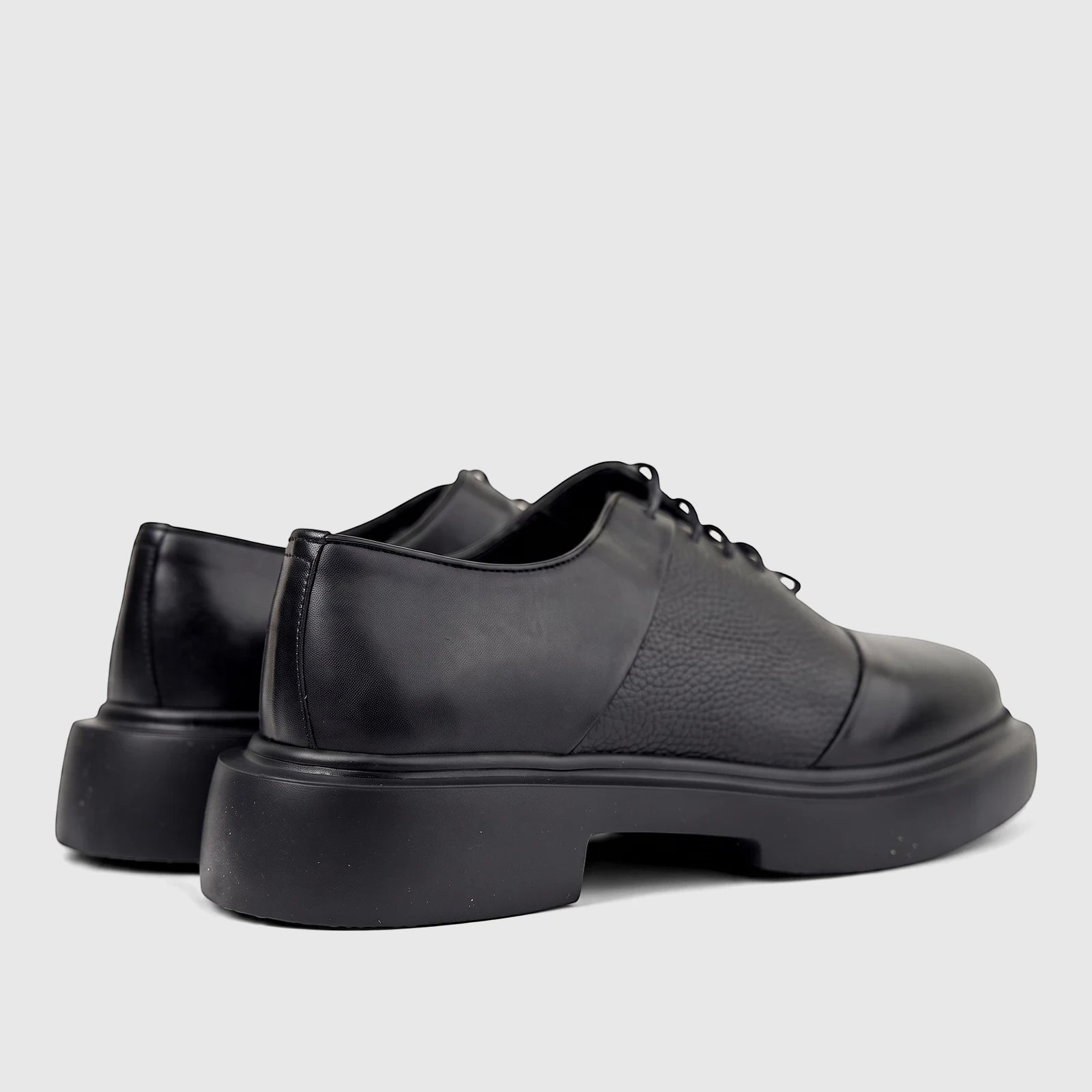 A pair of men's black leather lace-up shoes with chunky soles, viewed from the back-right showing the textured upper.