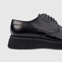 Close-up of a black patent leather men's formal shoe, featuring a hi-shine finish, chunky sole, and lace-up design.