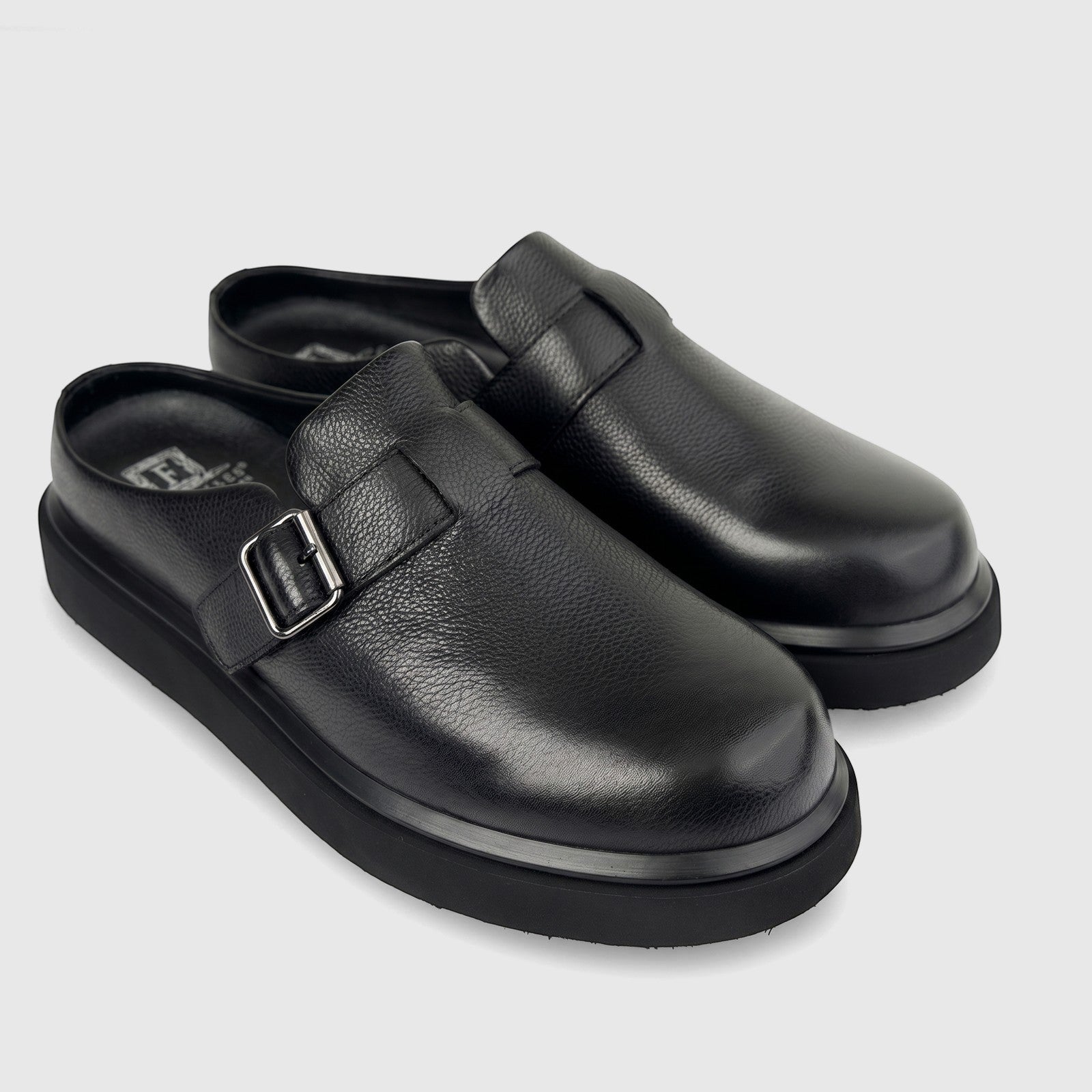 A pair of black Formales men's leather chunky sole mules with silver buckle details, viewed from the side on a white