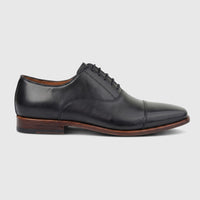 Black leather men's dress lace-up shoe with a cap toe and brown sole, viewed from a side profile on a white background.