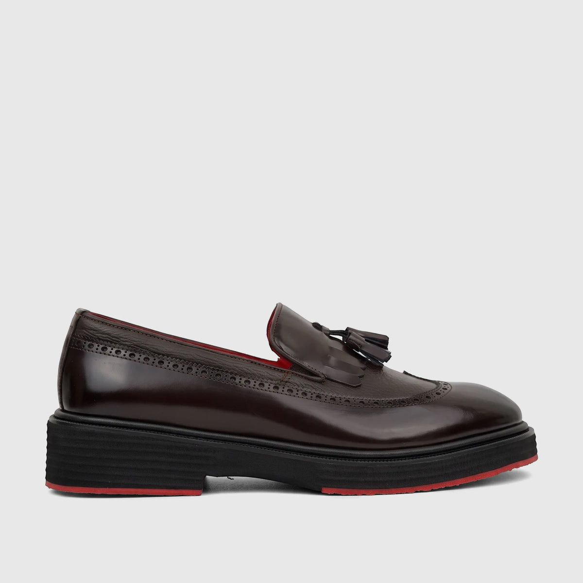 Dark brown leather unisex loafer with a chunky black sole, red trim, brogue detailing, and tassels, shown from a side view.