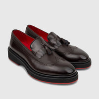 A pair of dark brown formal unisex leather chunky sole loafers with brogue details, tassels, fringe, and striking red