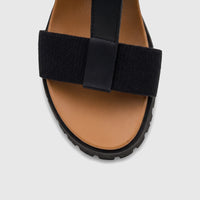 A close-up view of a black and tan HP Ladies Comfort Wedge Sandal with elastic and T-straps, and a thick black sole.