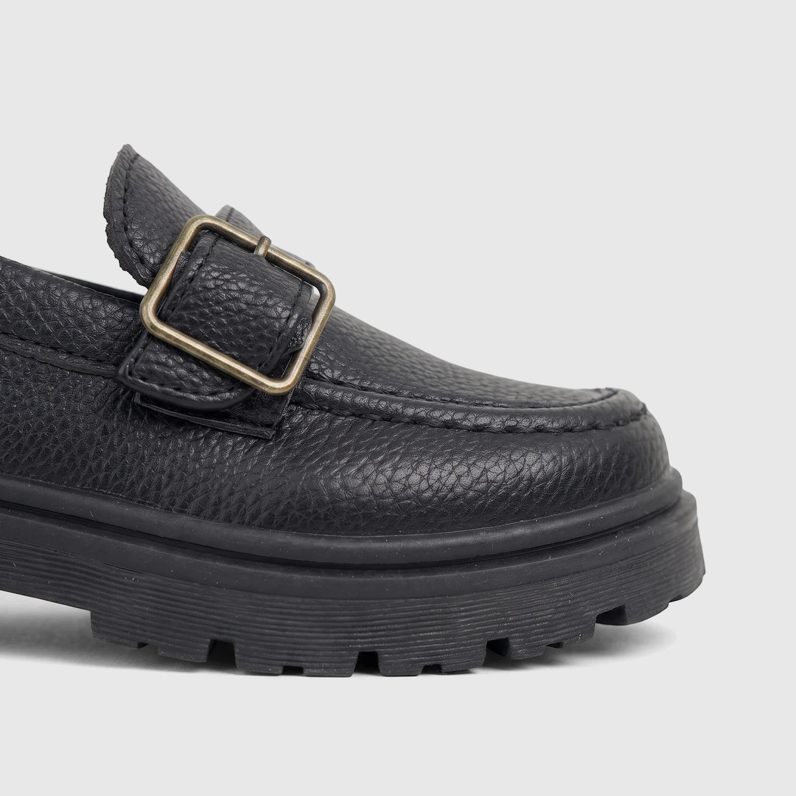 Close-up of a black textured leather chunky sole loafer with a bronze monk strap buckle on a white background.