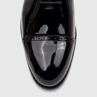 Men's black patent leather lace-up dress shoe, close-up of the polished toe cap with classic brogue detailing.