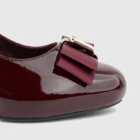 Close-up of a glossy wine red patent leather ladies' dress court shoe with a grosgrain ribbon bow and gold accent.