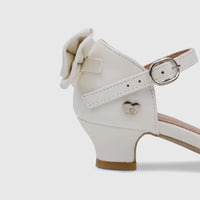 White PC girls' dress heel shoe with a bow, silver buckle on the ankle strap, and a heart emblem, viewed from the side.