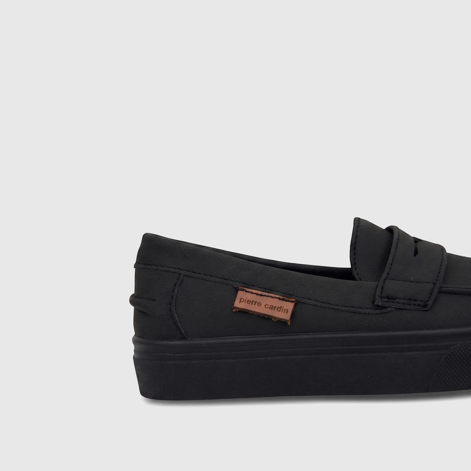 A black Pierre Cardin boys' casual slip-on shoe, featuring a textured upper and a brown logo tag, presented in a close-up