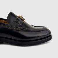 Close-up of a men's black leather Valentino slip-on loafer with a chunky sole and gold-tone 'V' logo hardware.