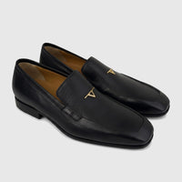 A pair of black Valentino Men's Leather Dress Slip On shoes with golden 'T' logo hardware and a square toe, displayed on a